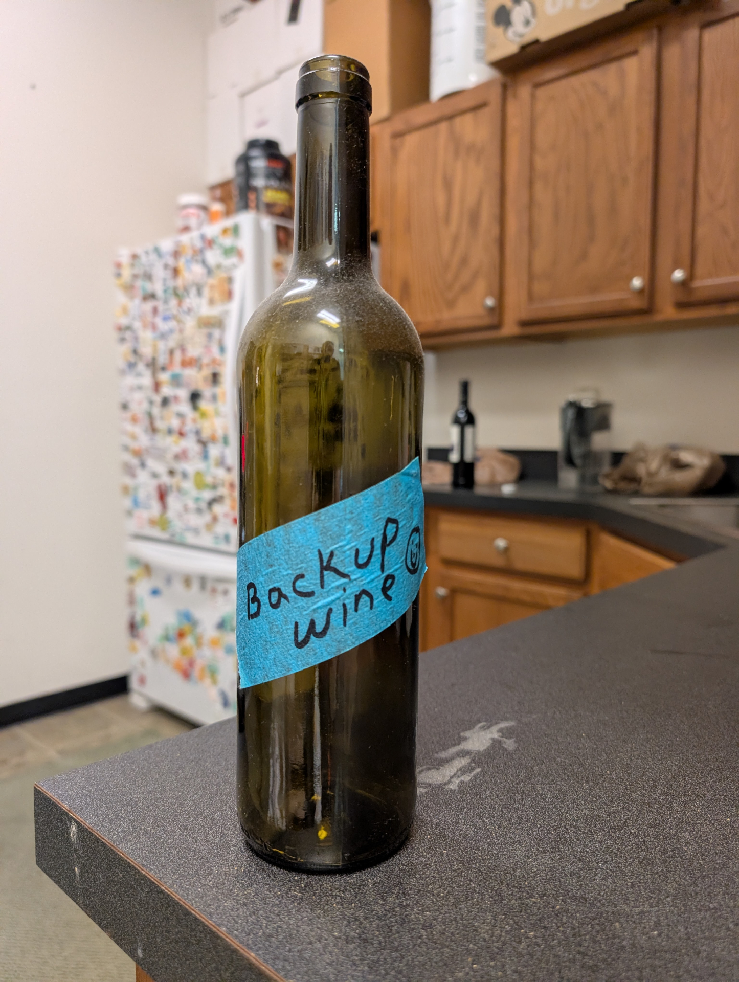 Empty green wine bottle on a kitchen counter with a strip of blue painter's tape across it reading 'Backup Wine' in black marker — the original handwritten label that started the brand
