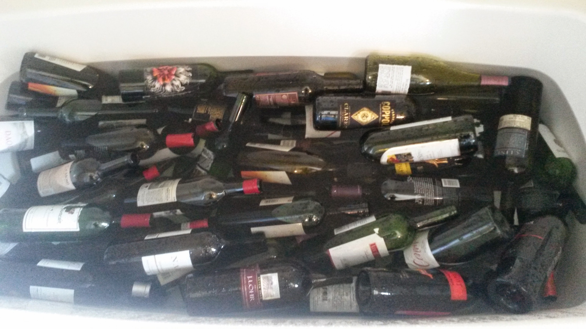 Looking down into a white bathtub packed with dozens of empty wine bottles laying on their sides and submerged, labels of various colors and shapes still attached, soaking in a few inches of water