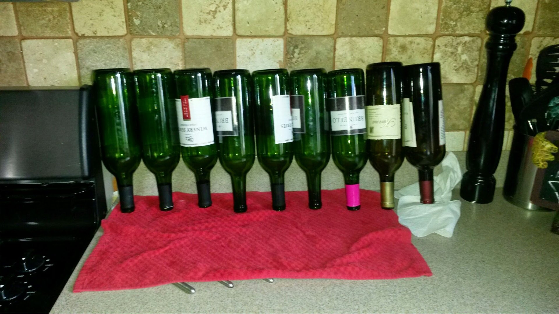 A row of about ten empty green wine bottles standing upside down to drain on a red kitchen towel next to a black stove, with a few labels still intact and a couple of colored foil capsules visible on the necks