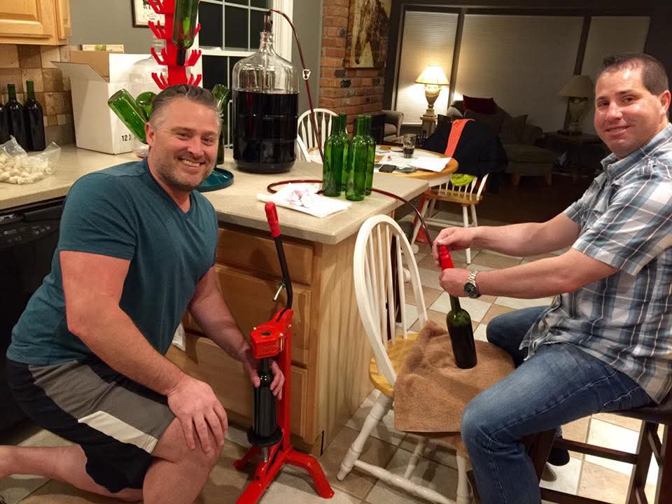 Russ (left, in a teal t-shirt) crouched next to the red floor corker grinning at the camera while Todd (right, in plaid) corks a green bottle by hand at the kitchen island, with a full carboy of dark wine and a bottle tree behind them