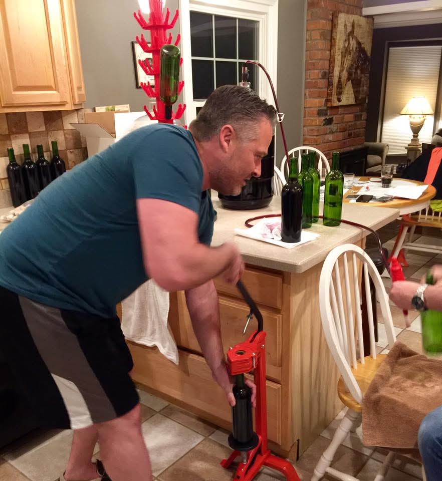 Russ leaning over a red floor corker on the kitchen tile, pressing a cork into a bottle of homemade wine on bottling day