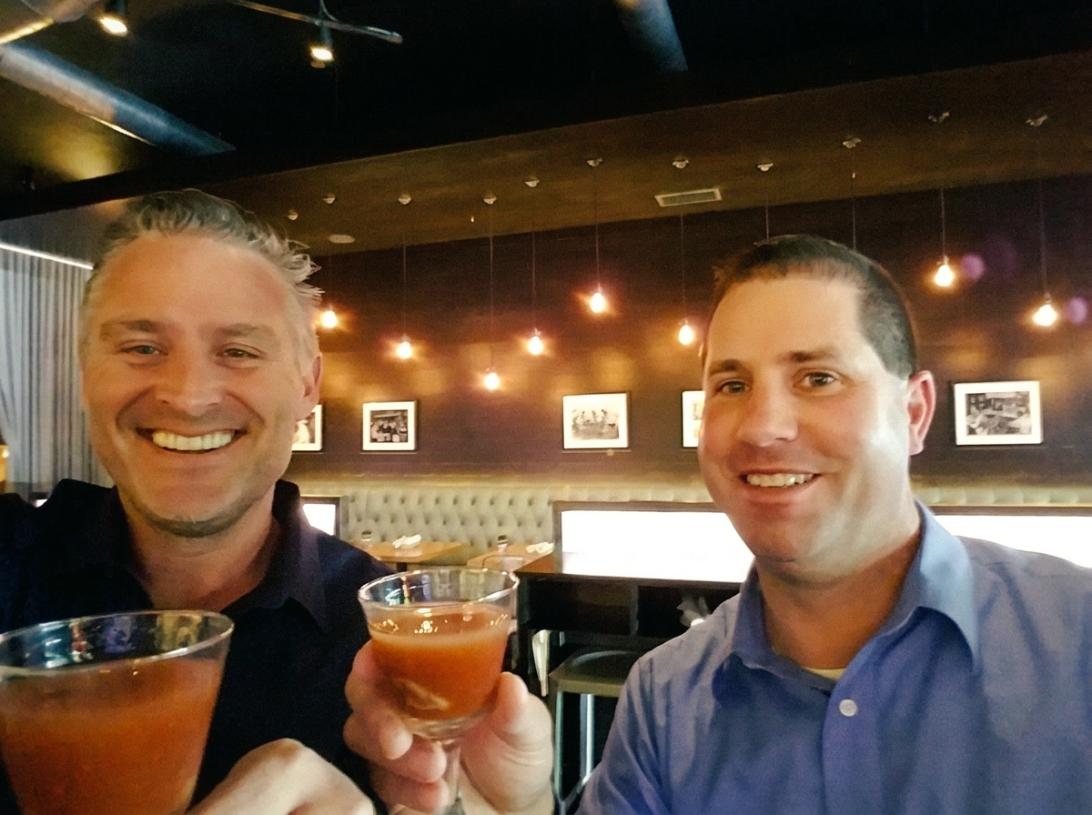 Todd and Russ toasting Bloody Marys at a happy hour, warm editorial parchment tone