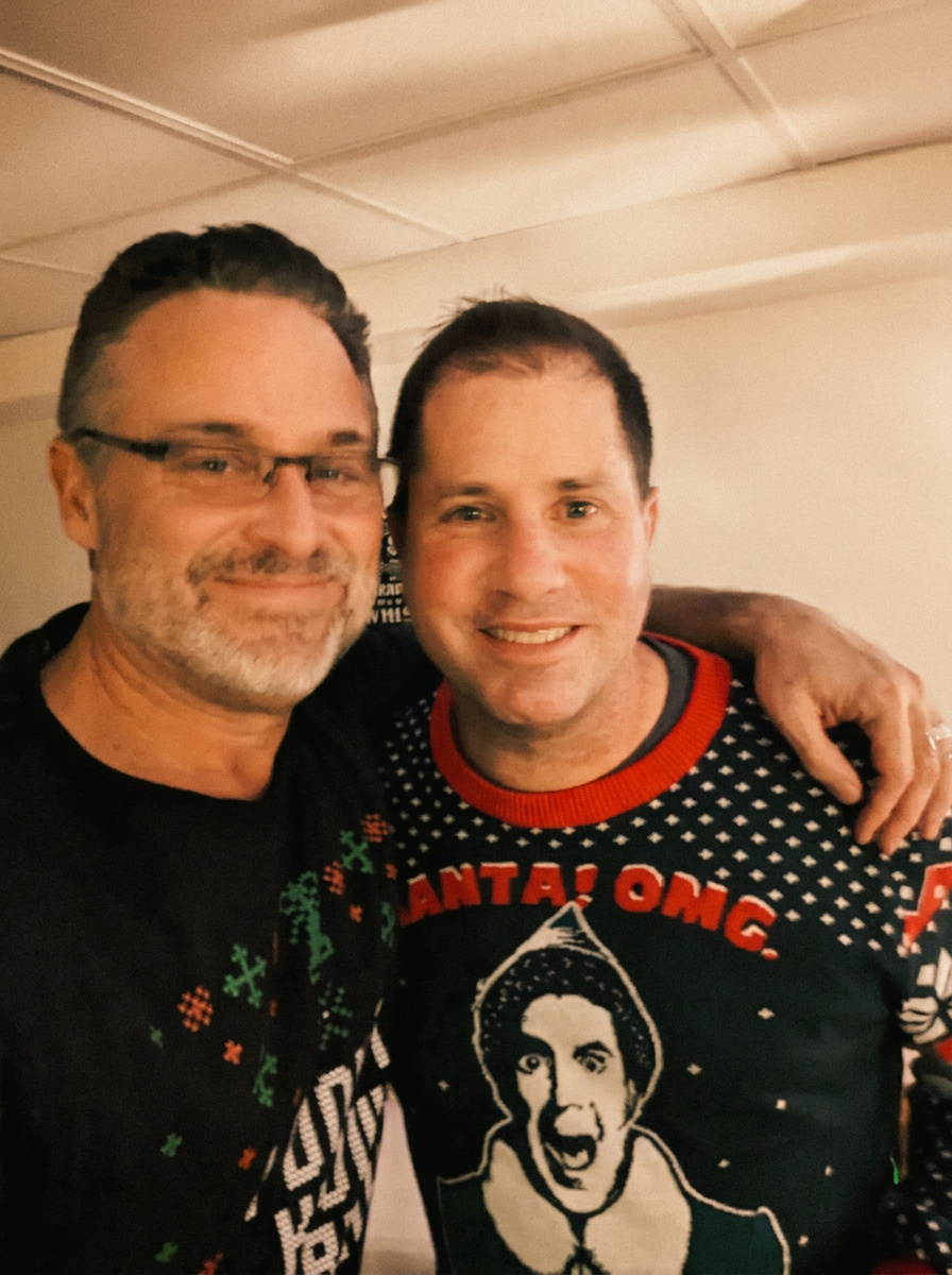 Todd (left, glasses) and Russ (right, in 'Santa! OMG!' Elf ugly Christmas sweater) at a holiday party, warm editorial parchment tone
