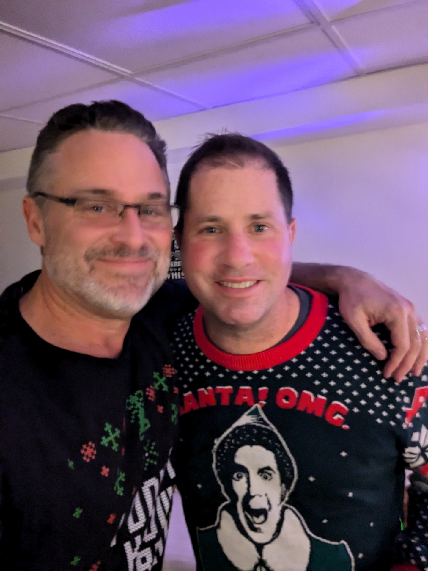 Todd (left, in glasses with a salt-and-pepper beard) and Russ (right, in a 'Santa! OMG!' Elf ugly Christmas sweater) smiling cheek-to-cheek at a holiday party