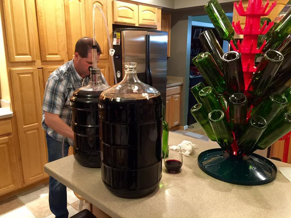 Todd leaning over two full carboys of dark homemade wine on the kitchen island, with a bottle tree of green bottles drying beside them
