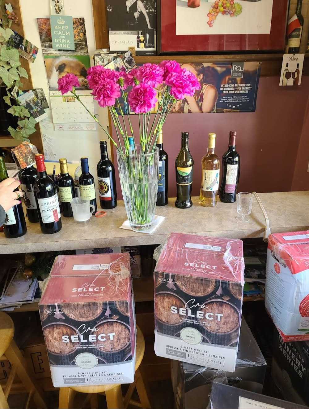 The tasting bar at Vine to Wine on bottling night: a vase of bright pink carnations surrounded by open bottles of red and white wine, two RJS Cru Select kit boxes on the floor below the counter