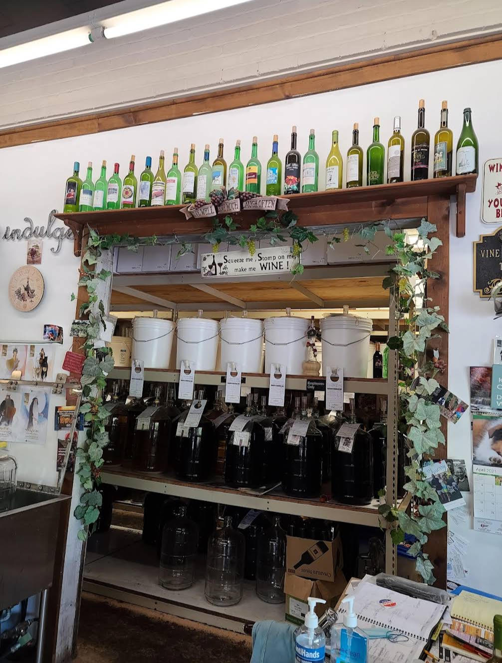 Rows of dark glass carboys full of fermenting red wine on the back shelves at Vine to Wine, with white plastic primary fermenters lined up on the shelf above and a 'Live Well, Love Much, Laugh Often' sign in the middle