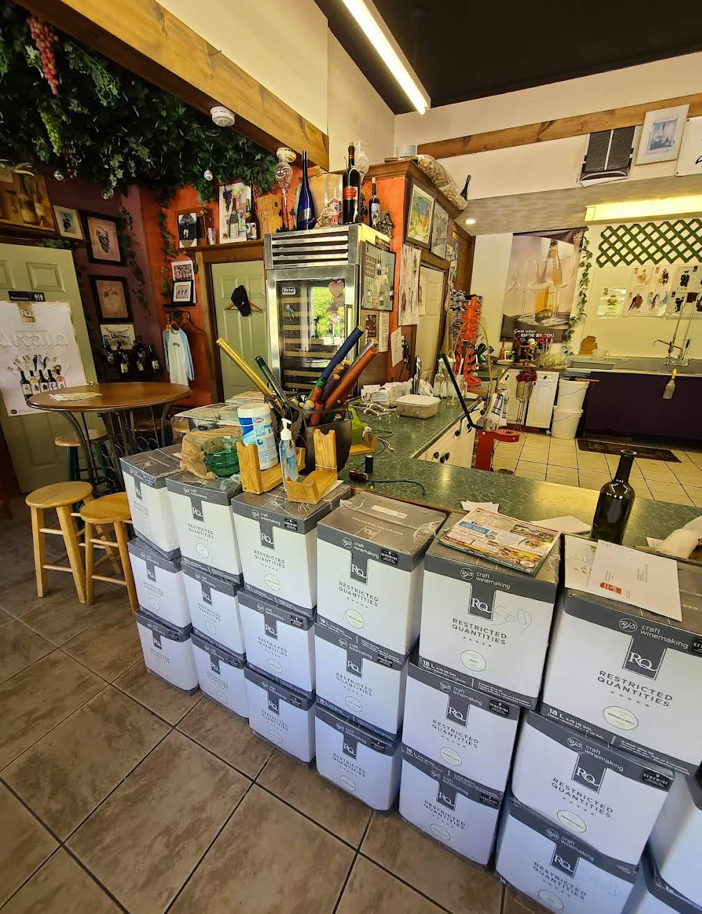 Stacks of RJS Craft Winemaking Restricted Quantities kit boxes piled chest-high in the front of the Vine to Wine shop, next to the tasting bar with stools