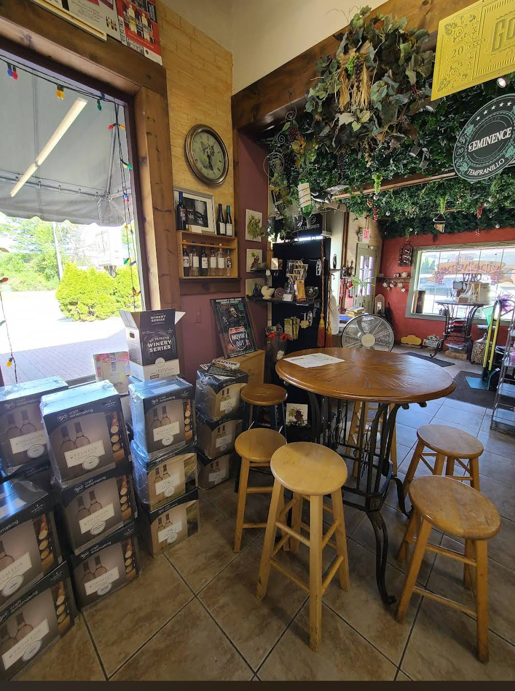 The Vine to Wine tasting corner: a round high-top table ringed with wooden barstools, dried hops and grape vines hanging overhead, kit boxes stacked alongside, the front roll-up door open to the parking lot